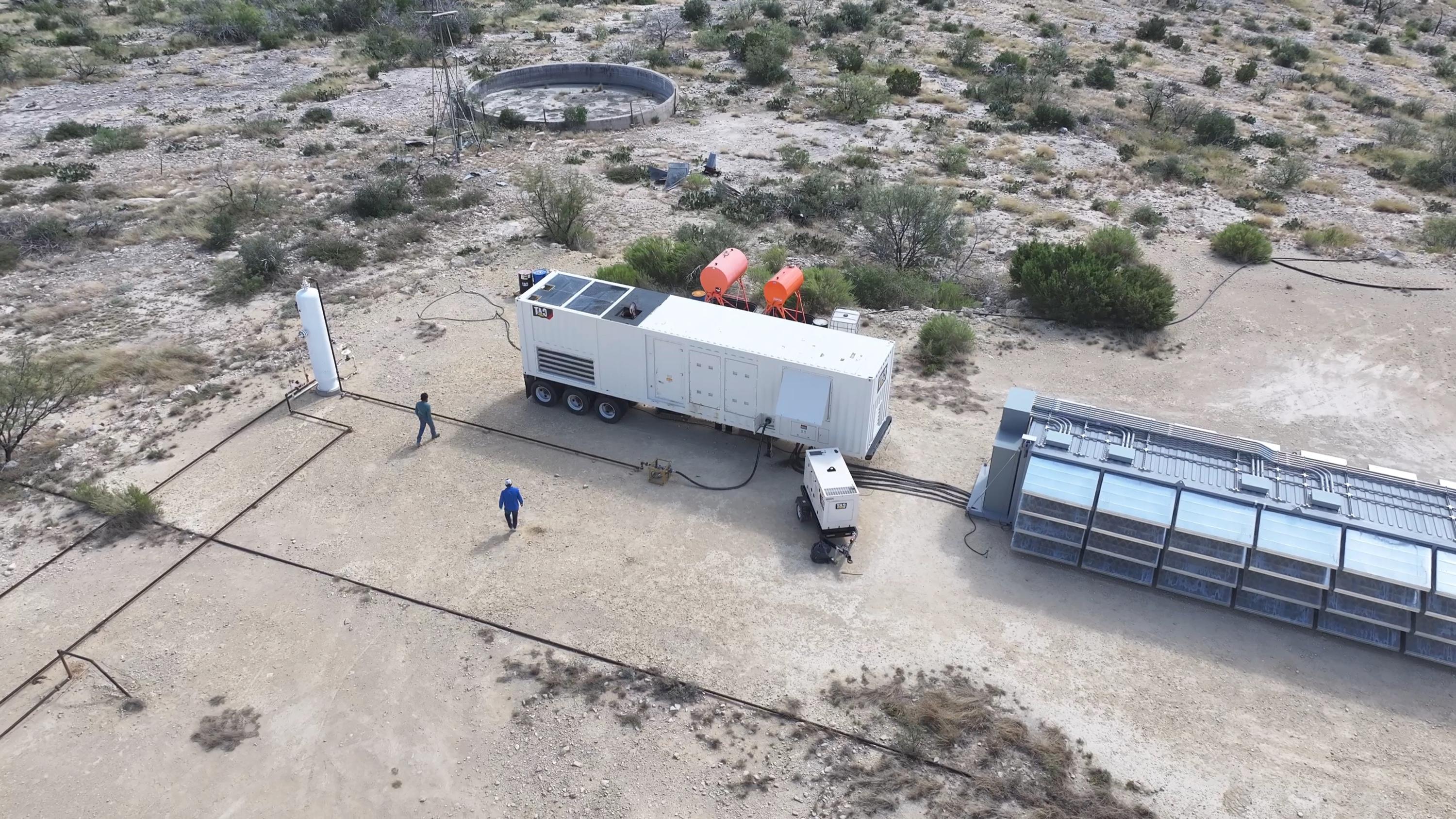 Aerial view of a remote site: a containerized power module on a trailer with radiator units on its roof, a smaller generator alongside, heavy cabling running to a long row of open-top immersion-cooled computing tanks, on cleared desert ground.
