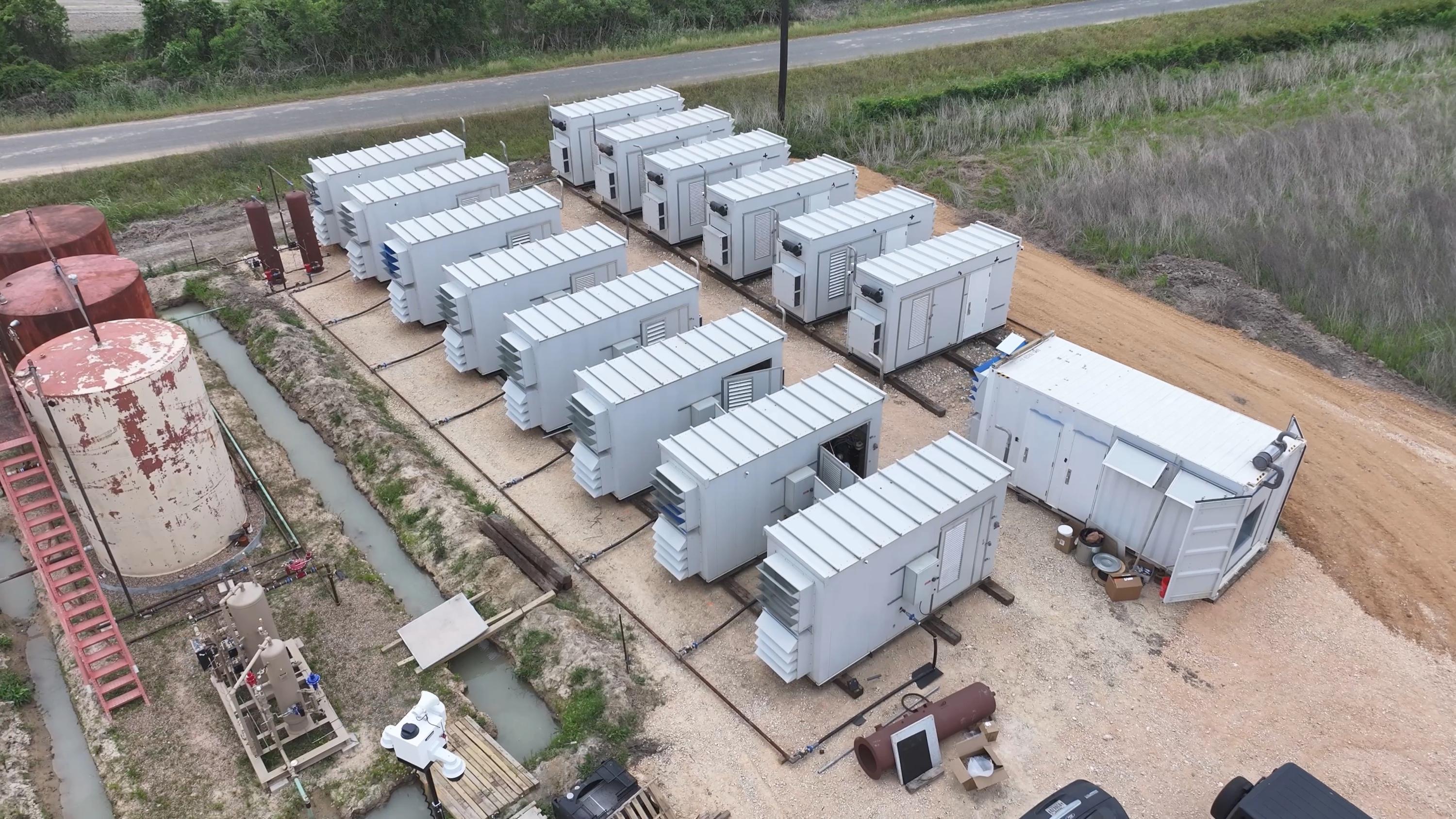 Aerial view of a modular deployment: rows of modular units on a graded pad beside a control container, with production tanks and wellsite equipment to one side and an access road running past the site.