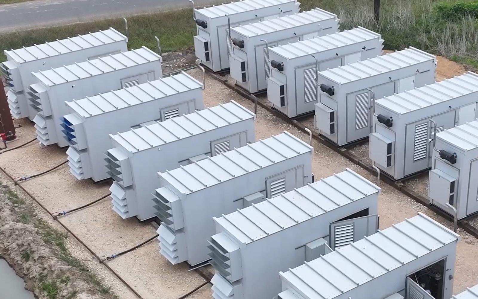 Aerial view of modular units arranged in rows on a prepared pad, each with side louvres and roof panels, connected by ground-level pipework and cabling.