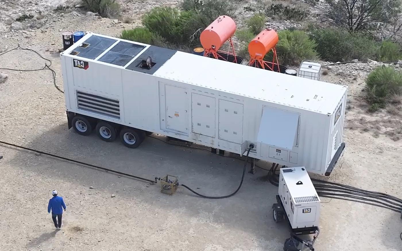 A containerized power module on a trailer with roof-mounted radiators and a side air intake, a smaller generator set beside it, and bundled cables running out across the ground.