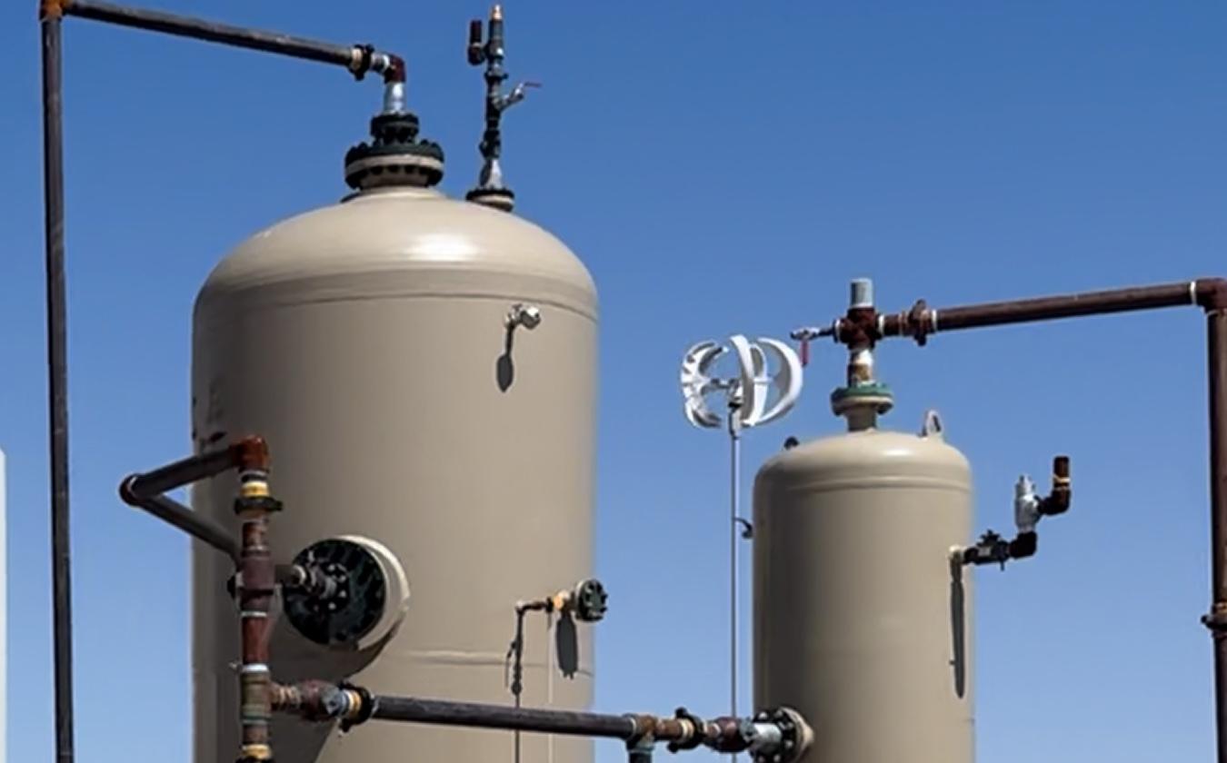 Ground-level view of a gas processing site: a row of storage tanks with an access stairway, two vertical separator vessels, connecting pipework and a control cabinet, on open desert ground.
