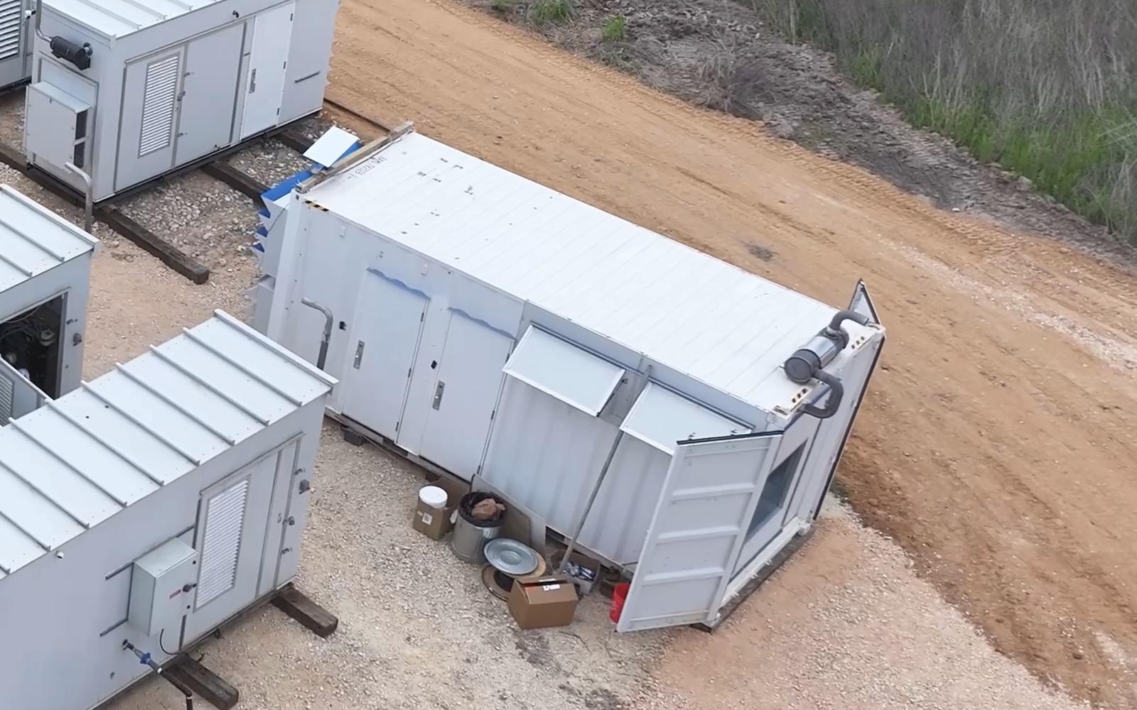 Aerial view of a site under build-out: rows of modular units on a graded pad beside a control container, with an access track running along the edge and materials laid down nearby.
